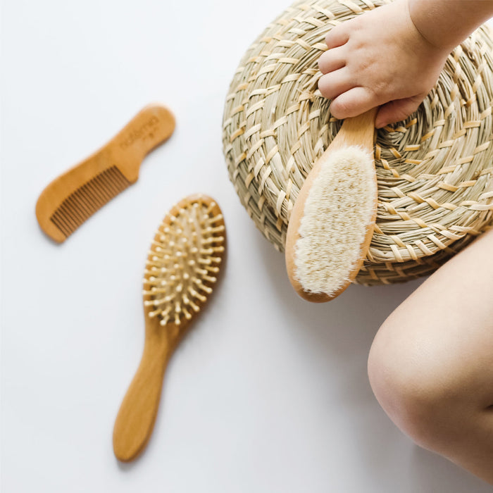 A child holds a woven basket topped with the Natemia Wooden Baby Hair Brush from the Hair Brush & Comb Set. Nearby are another brush with rounded bristles and a wooden comb, essential baby grooming accessories. The plain white surface serves as the backdrop.