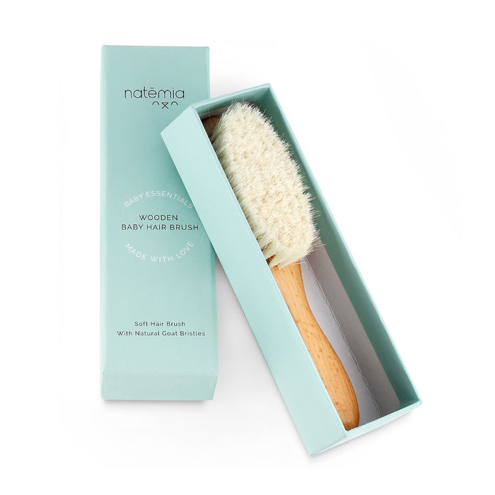 The Natemia Wooden Baby Hair Brush features soft natural goat bristles and comes in an elegant open teal box. Highlighted as a cradle cap brush, the packaging emphasizes its gentle softness and status as a baby essential, made with love.