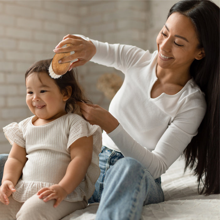 A woman in a white long-sleeve shirt gently brushes a smiling toddlers hair with the Natemia Wooden Baby Hair Brush. The toddler, in a beige short-sleeve shirt, sits on a bed in softly lit surroundings with an exposed brick wall that adds rustic charm.