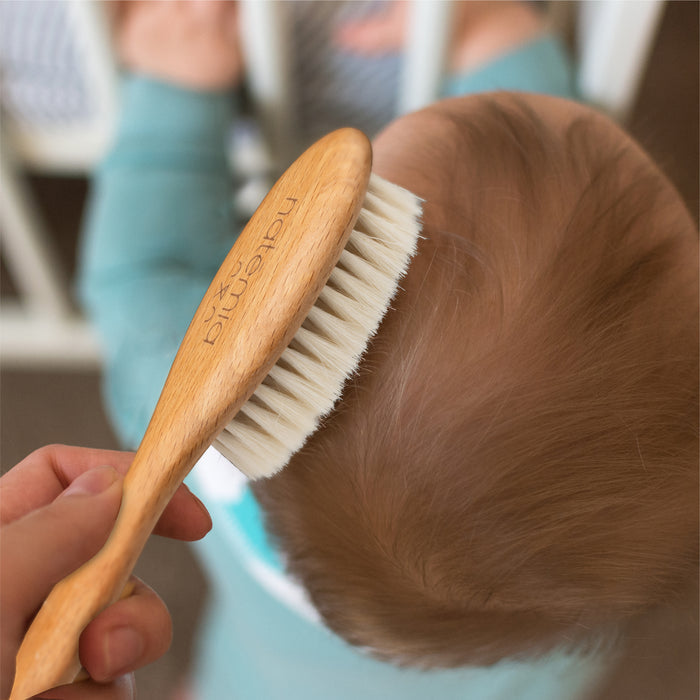 A hand gently brushes a babys hair with the Natemia Wooden Baby Hair Brush. The baby, wearing a teal outfit, turns away against the blurred backdrop of white crib bars.