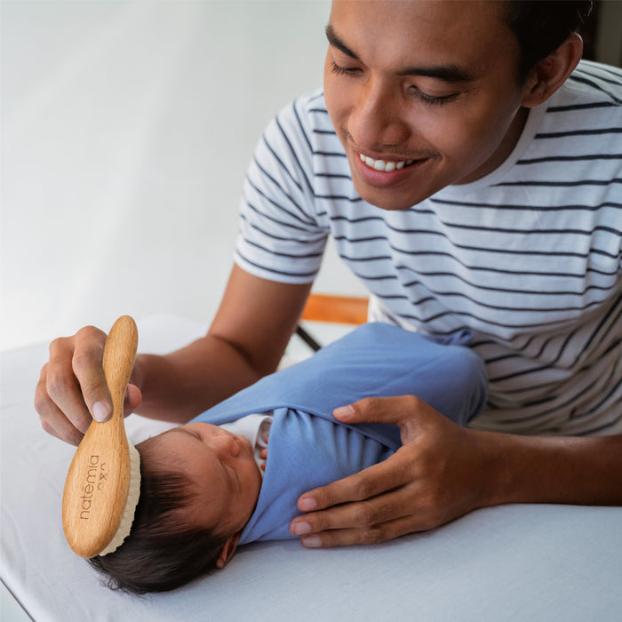 A man in a striped shirt smiles as he gently uses Natemias Wooden Baby Hair Brush to smooth the hair of a swaddled baby. The infant, wrapped in a blue blanket, lies peacefully on a white surface as natural bristles work their magic.
