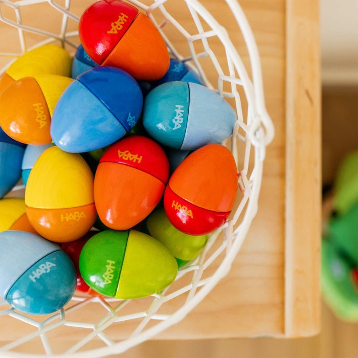 A white wire basket is filled with colorful, split egg-shaped HABA Wooden Musical Eggs in blue, orange, red, green, and yellow. They display HABA and rest cheerfully on the wooden surface.
