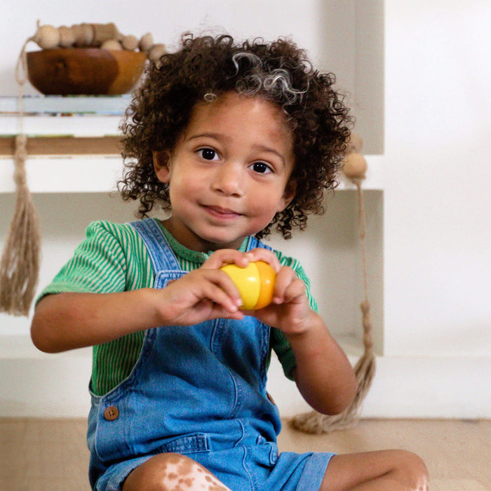 A curly-haired child with a vitiligo patch on one leg smiles indoors, holding a yellow bell pepper. Theyre dressed in a green striped shirt and blue denim overalls. Wooden Musical Eggs by HABA are displayed on shelves with other decor items in the background.