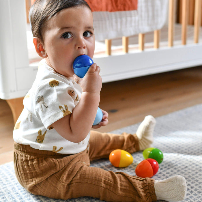 A toddler in a white animal-print shirt and brown pants sits on a rug, holding a blue toy egg in their mouth. They are surrounded by colorful HABA Wooden Musical Eggs. In the background, a crib highlights the love for sustainable toys.