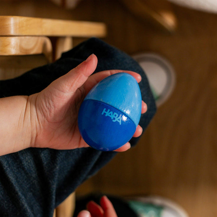A child in dark pants grasps a blue, egg-shaped Wooden Musical Egg by HABA while sitting on a wooden chair, surrounded by shoes and wooden flooring—a nod to sustainable toys.