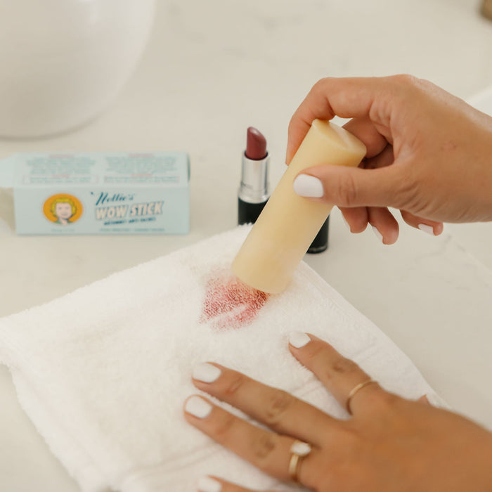 A hand with white nail polish and gold rings rubs Nellie’s WOW Stick Stain Remover, a natural stain remover, on a lipstick stain on a white cloth. A lipstick and the blue WOW Stick box are visible in the background.