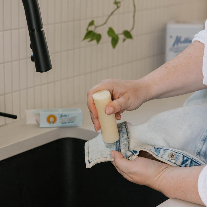Someone rubs Nellie's WOW Stick Stain Remover on the cuff of light-wash denim jeans over a kitchen sink, with a plant and other green cleaning products visible in the background.