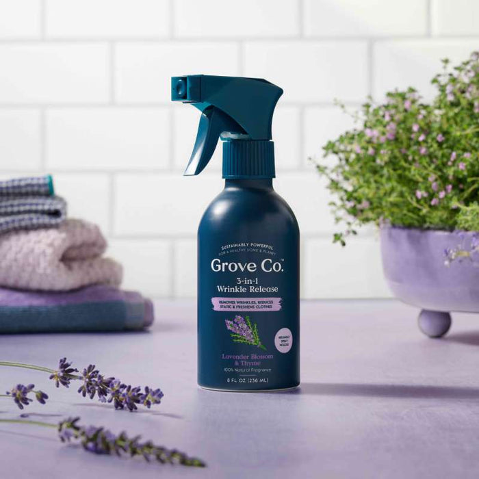 A bottle of Grove Co.s 3-in-1 Wrinkle Release rests on a lavender surface, complemented by fresh lavender sprigs and folded towels in the background for a clean, calm ambiance.