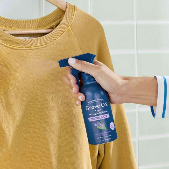 A hand sprays Grove Co. 3-in-1 Wrinkle Release with natural fragrance on a mustard shirt on a wooden hanger. The blue bottle, labeled with lavender scent, stands out against the tiled background, highlighting its plastic-free packaging.
