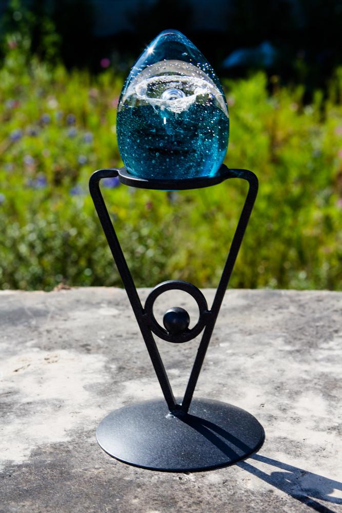 Wrought iron triangle stand holding a blue glass orb outdoors on a stone surface.