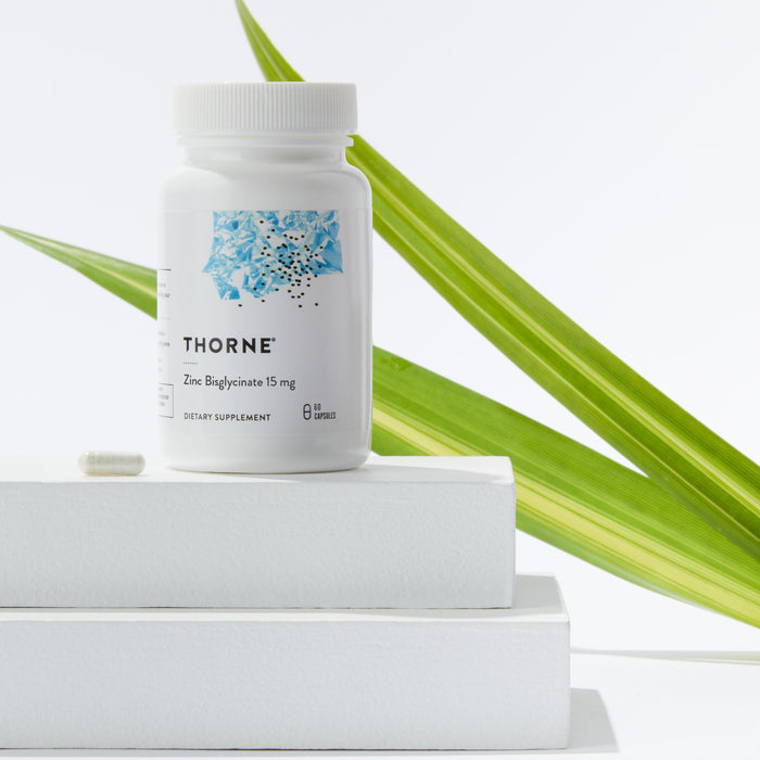 A Thorne Zinc Bisglycinate 15mg supplement bottle is on two stacked white platforms with one capsule in front, and a green leaf in the background symbolizing its support for immunity and reproductive health.