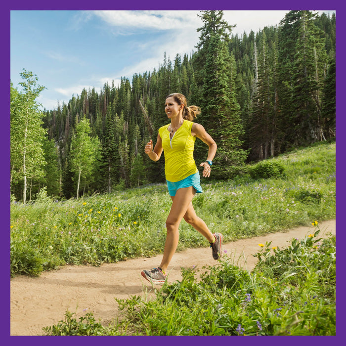 A woman jogs on a dirt trail in a yellow tank top and blue shorts, surrounded by lush greenery and pine trees, under a clear sky. She stays focused and active with Zyflamend™ Herbal Pain Relief by New Chapter, enjoying natural inflammation relief in the serene setting.